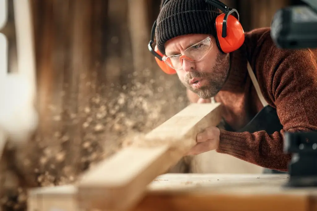 Mann hobelt konzentriert ein Holzbrett in der Werkstatt – Symbolbild für körperliche Arbeit und Absicherung durch die HUK-COBURG Berufsunfähigkeitsversicherung.